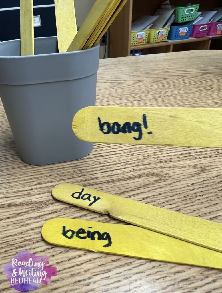Pictures of yellow popsicle sticks with words "day" and "being" on them, on the table , and popsicle stick with word "bang!" being held up